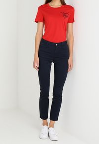 Rode katoenen T-shirt met korte mouwen en een gedrukt logo, gecombineerd met donkerblauwe slim-fit jeans en witte sneakers. Gladde textuur.