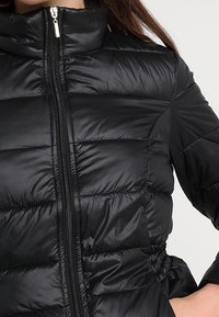 Svart puffjacka med hög krage, dragkedjestängning och kviltad textur; har elastiska detaljer vid midjan och ärmarna.