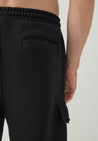 Sorte sweatpants med struktureret stof, elastisk talje og en sidelomme med klaff. Glat, tætsiddende design med minimal pynt.