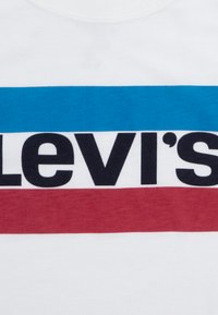 Baltas kokvilnas T-krekls ar melnu, izteiksmīgu "Levi's" logotipu, virs un zem logotipa horizontālas zilas un sarkanas svītras.