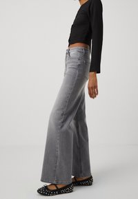Femme portant un jean gris taille haute à jambes larges, un crop top noir à manches longues et des ballerines noires cloutées, debout de profil.