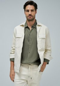Helles beiges Jacket mit zwei Brusttaschen, über einem olivgrünen Button-Up-Hemd getragen. Eine bequeme Cremefarbene Hose mit Kordelzug rundet das Outfit ab.