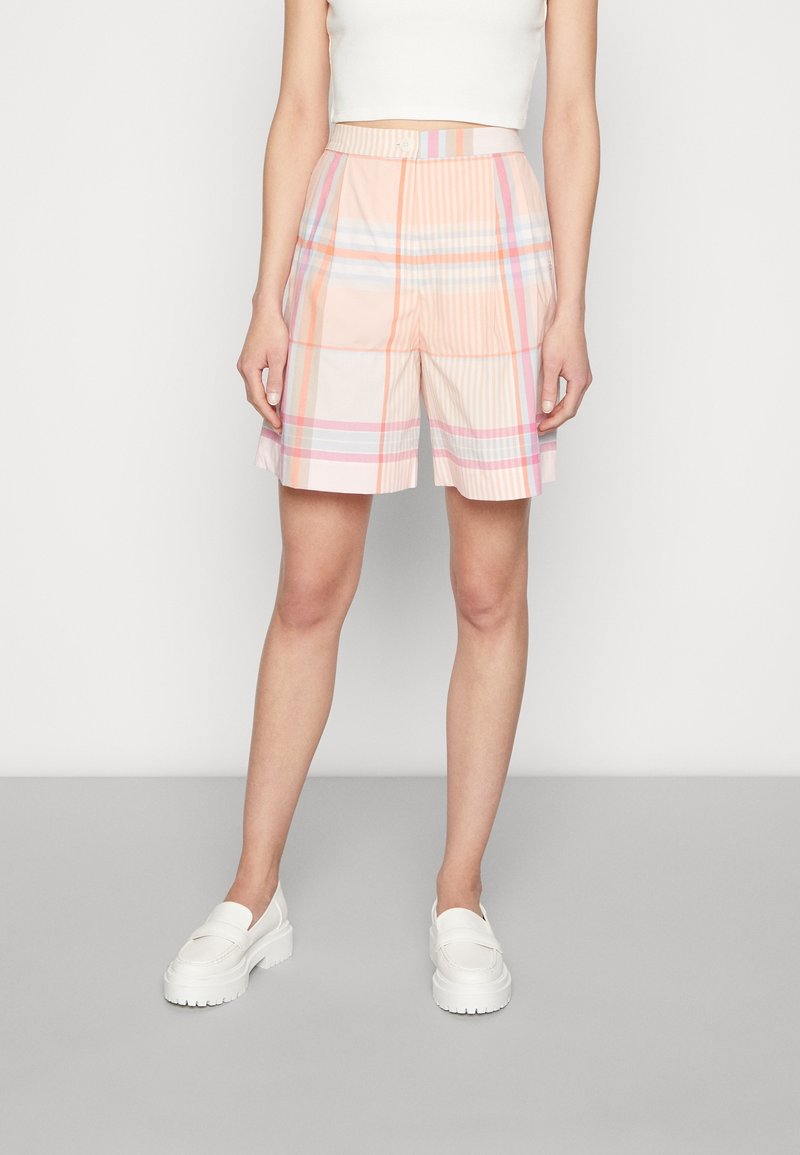 Femme portant un short à carreaux pastel taille haute avec des plis devant, assorti à un top court blanc et des mocassins blancs épais, debout sur un sol gris.