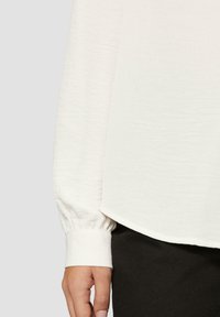 Biała bluzka z teksturowanej tkaniny, z marszczonym mankietem i zaokrąglonym dołem. Noszona na czarne spodnie, minimalistyczny design.
