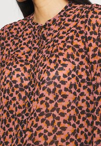 Långärmad blus med en rosa bakgrund och svart och orange blommönster, mjukt tyg och en knappslå med subtila veck.