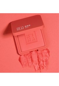 Compact de blush corail avec logo en relief, de forme rectangulaire et surface mate, accompagné d'un échantillon du produit sur un fond assorti.