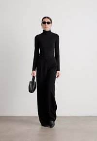 Maglione a collo alto fitted nero abbinato a pantaloni neri a vita alta, occhiali da sole neri e una borsa nera strutturata, in un ambiente minimalista.