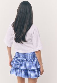 Donna con capelli lunghi e scuri che indossa una camicia bianca e una gonna a balze a quadretti blu e bianchi, in piedi contro uno sfondo chiaro e uniforme.