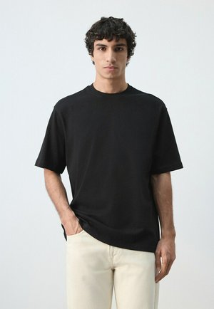 Young man with curly dark hair wears a plain black short-sleeve shirt and light beige pants, standing against a plain light background.