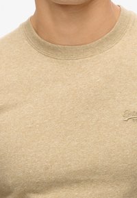 Beige short-sleeve t-shirt made of soft, textured fabric. Features a round neck and embroidered logo on the chest. Simple design.