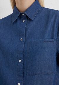 Dunkelblaues Denimhemd mit durchgehender Knopfleiste, spitzem Kragen und einer einzelnen Tasche auf der linken Brust. Dezent aufgesticktes Markenlogo.