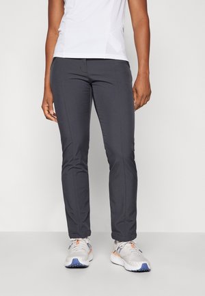 Osoba ubrana w czarne, dopasowane spodnie, białe sportowe buty z niebieskimi podeszwami oraz białą koszulę z krótkim rękawem, stojąca na szarej podłodze.