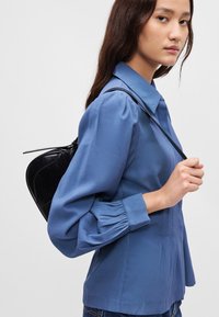 Μπλε πουκάμισο με μακριά μανίκια, κουμπωτό γιακά, πλισέ μανσέτες και χαλαρή εφαρμογή. Το μοντέλο φοράει μια μαύρη, υφασμάτινη τσάντα πλάτης.