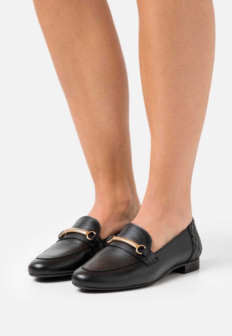 Mocassins en cuir noir avec détail en métal doré, portés pieds nus, dotés d’un petit talon et d’une texture matelassée à l’arrière du talon.