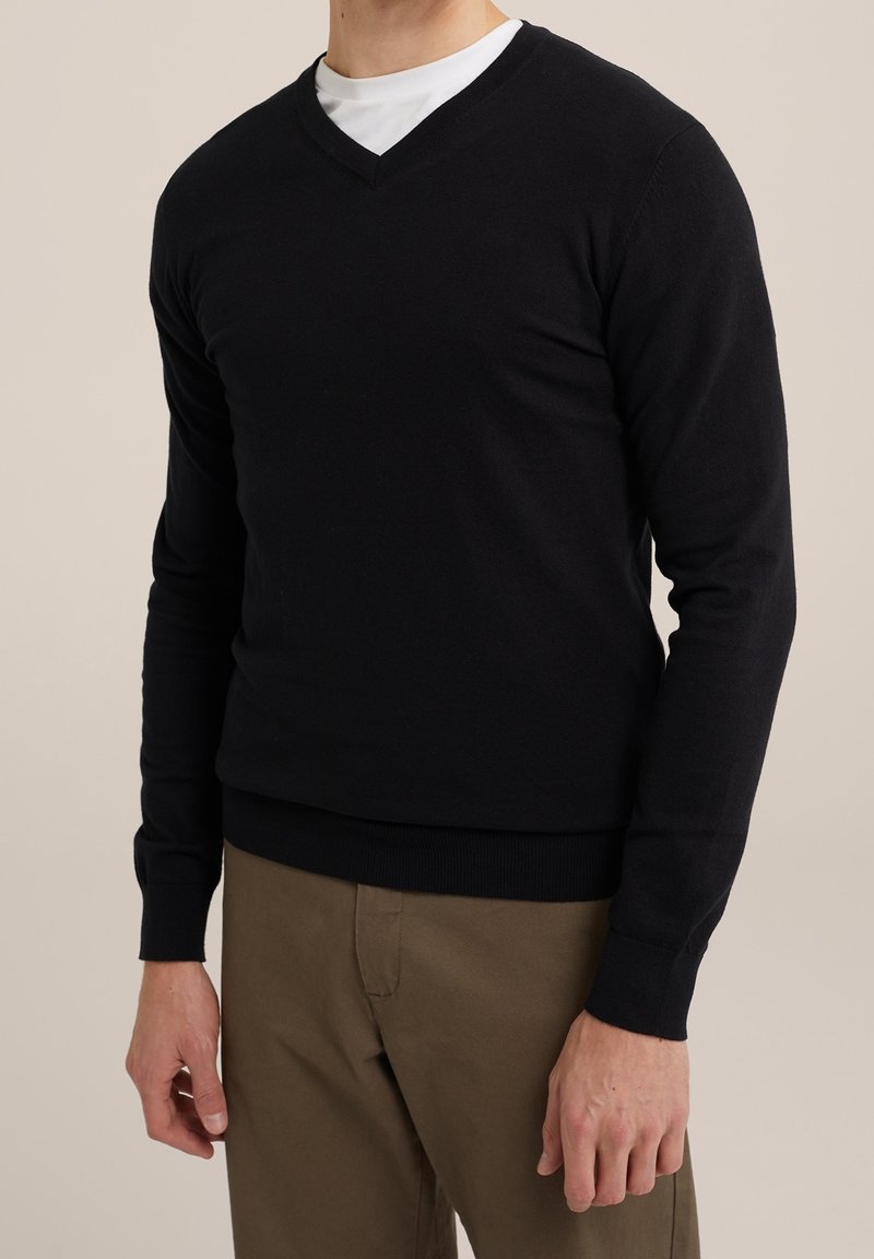 Musta v-kaula-aukollinen neulepaita istuvassa leikkauksessa, pitkät hihat, resorit helmassa ja hihansuuissa, valmistettu pehmeästä materiaalista, pidetty valkoisen paidan päällä ja yhdistetty vaaleanruskeisiin housuihin.