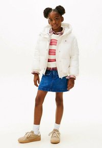 Balta pūkaina jaka ar kapuci, kopā ar svītrainu džemperi un džinsa svārkiem. Apģērbu papildina brūnas krāsas sporta apavi un baltas zeķes.