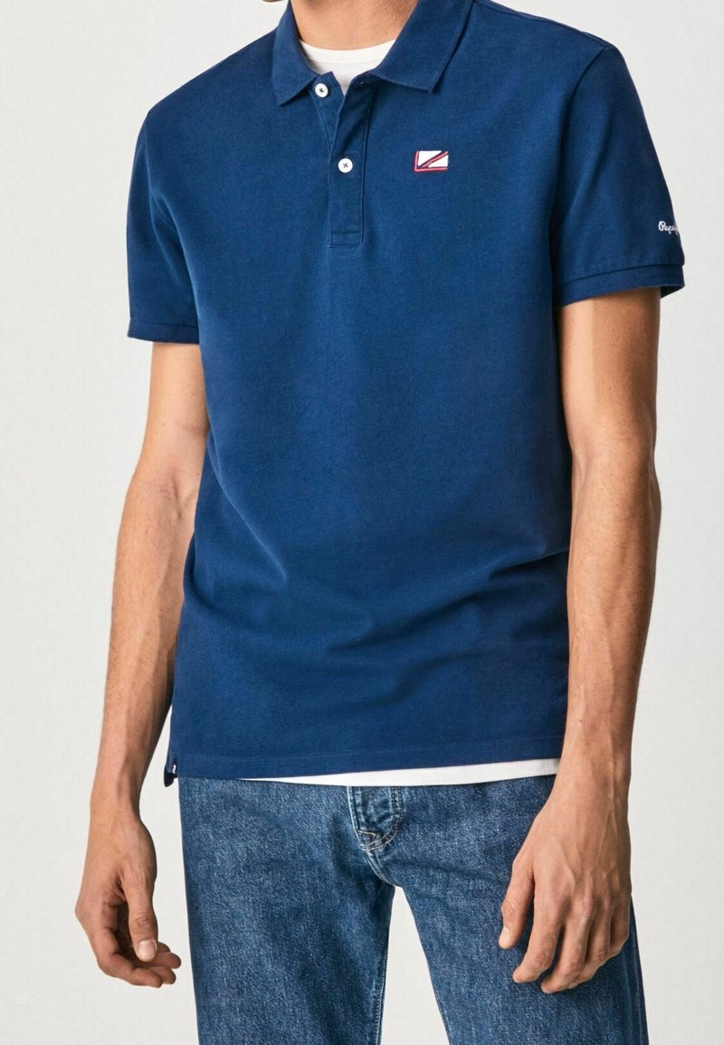 Homme portant un polo à manches courtes bleu marine avec un petit logo sur la poitrine et un jean bleu clair, se tenant avec les bras détendus le long du corps.