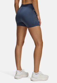 Marinefarbene Kompressionsshorts mit hohem Bund, ausgestattet mit einem kleinen Logo-Patch, kombiniert mit weißen Sportschuhen und strukturiertem Stoff.