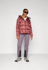 Una persona con el cabello trenzado lleva una chaqueta de abrigo burdeos, pantalones grises y gafas de sol, de pie contra un fondo liso.