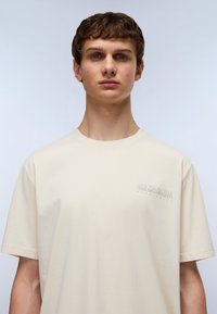 T-shirt en coton beige à manches courtes, col rond, avec un petit logo imprimé discret sur la poitrine gauche. Texture lisse.
