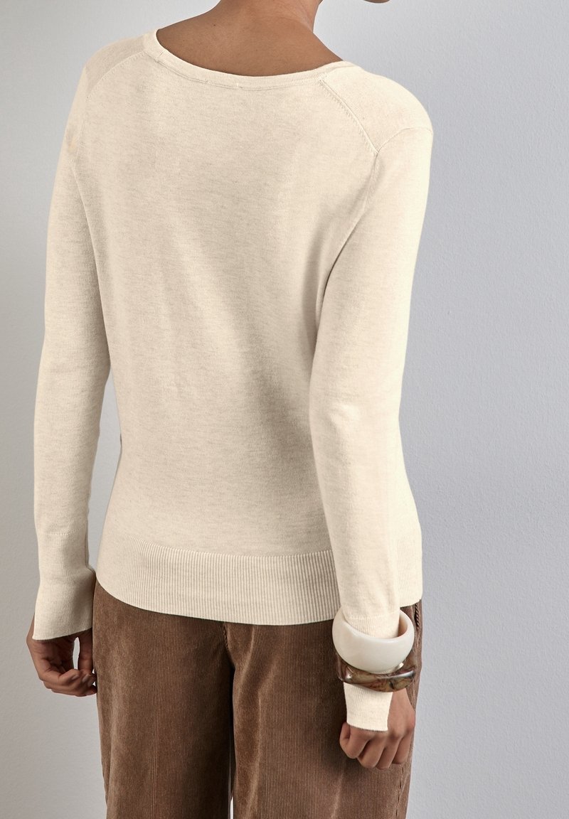 Personne portant un pull crème à manches longues et un pantalon en velours côtelé marron, avec des bracelets blancs et bruns au poignet gauche, vue de dos.