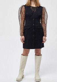 Schwarzes gestreiftes Kleid mit taillierter Oberseite und Knöpfen, das über einem transparenten Langarmoberteil mit Leopardenmuster getragen wird. Cremefarbene knielange Stiefel.