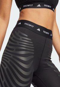 Černé legíny s širokým pasem a logem "TECHFIT". Na boku mají lesklý, vlnitý vzor, který dodává texturu a detaily.