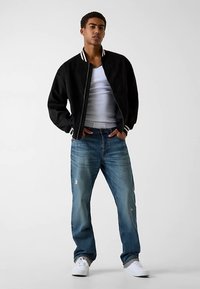 Schwarze Bomberjacke mit weiß gestreiftem Kragen, kombiniert mit hellblauen, verwaschenen Jeans und weißen Turnschuhen. Einfaches, lässiges Outfit.