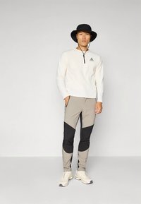 Lys langermet skjorte med halv glidelås, grå og svarte fargeblokkert smale bukser, og en svart bøttehatt med en liten logo.