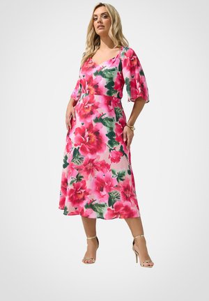 Kvinde iført en pink midi-kjole med store røde blomster og grønne blade, kombineret med gyldne højhælede sandaler og et armbånd.