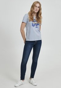 Camiseta de algodón azul claro con un diseño gráfico en azul y amarillo, combinada con vaqueros ajustados azul oscuro y zapatillas blancas.