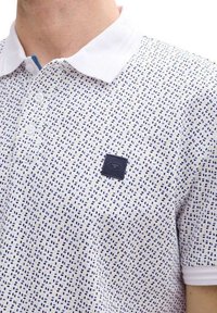 Chemise polo blanche avec une texture à motifs comportant des pois bleus et navy. Comprend un col contrasté et un patch de marque noir sur la poitrine.