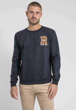 Homme portant un sweat-shirt bleu marine avec un écusson orange en forme de lettre "R" varsity, pantalon beige, se tenant devant un fond clair uni.