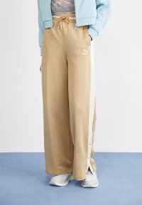 Pantalones de chándal anchos beige con bolsillos frontales y rayas blancas en los lados. La tela es suave, con una cintura ajustable mediante cordón y botones a presión cerca del dobladillo.