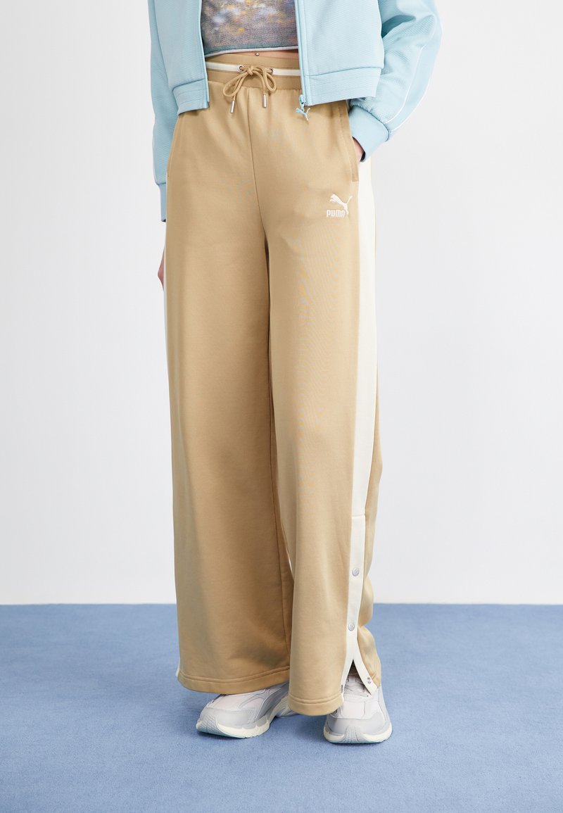 Pantalones de chándal anchos beige con bolsillos frontales y rayas blancas en los lados. La tela es suave, con una cintura ajustable mediante cordón y botones a presión cerca del dobladillo.