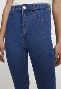 Gros plan d'une personne portant un jean en denim bleu taille haute avec un bouton et une fermeture éclair à l'avant, et un haut blanc rentré à l'intérieur.