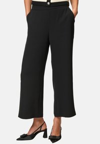 Pantalons noirs à jambe large, fabriqués en tissu lisse. Comprend des poches avant et une taille avec un bouton décoratif. Associés à des talons slingback noirs.