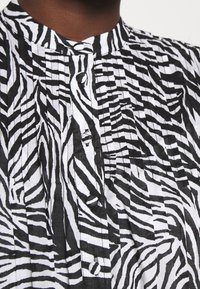 Černobílá košile s pruhy zebra, stojáčkovým límcem, zapínáním na knoflíky a sklady. Měkká tkanina a výrazné detaily vzoru.