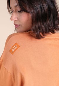T-shirt en coton orange avec une coupe décontractée, arborant une étiquette Oxbow cousue sur l'épaule et un fini de tissu lisse et texturé.