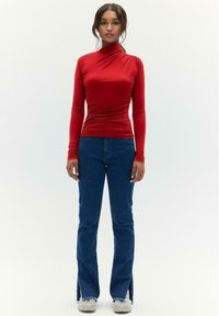Rode longsleeve top met een hoge kraag en geplooide textuur, gecombineerd met blauwe uitlopende jeans met zijsplitten en rafelige lof.