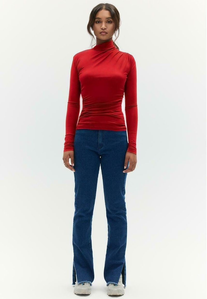 Rode longsleeve top met een hoge kraag en geplooide textuur, gecombineerd met blauwe uitlopende jeans met zijsplitten en rafelige lof.