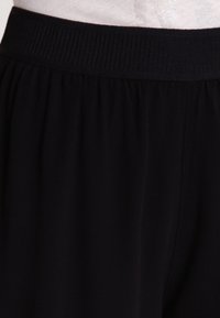 Pantalon noir en tissu doux et léger avec une ceinture côtelée et une coupe ample et fluide. Aucun motif ni détail métallique visible.