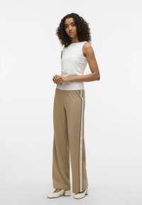 Witte mouwloze top gecombineerd met beige wijde broek met zwarte zijstrepen. Model staat voor een neutrale achtergrond.