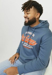 Blå bomulds hoodie med orange grafisk tekst og logo på forsiden. Har en hætte med snore og ribbede manchetter. Casual fit.