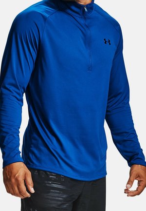 Mann trägt ein blaues, langärmeliges Sportoberteil mit Reißverschluss, schwarzem Under Armour-Logo und schwarzen, gemusterten Shorts vor weißem Hintergrund.