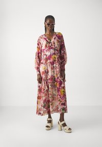 Bloemrijke maxi-jurk met gerafelde randen in tinten roze, geel en groen; lange mouwen. Gecombineerd met witte plateau-sandalen.