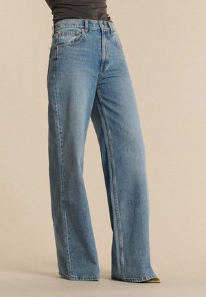 Osoba ubrana w niebieskie dżinsy o wysokim stanie i szerokich nogawkach, połączone z czarnym topem, stojąca na tle jednolitego beżowego tła.