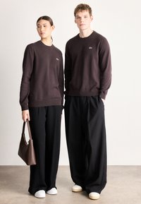 Camisolas de malha castanho escuro com golas redondas, combinadas com calças largas pretas. A mulher segura uma mala castanha; ambos usam sapatos brancos.