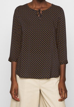 Czarna bluzka w pomarańczowe groszki, z rękawami do ćwiartki i małym wiązaniem z przodu, noszona z beżowymi spodniami.