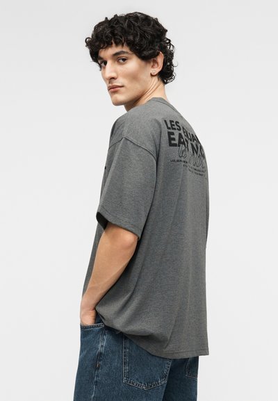 Tânăr cu păr creț, purtând un tricou gri cu imprimeu și blugi albaștri, uitându-se peste umăr cu o mână în buzunar.